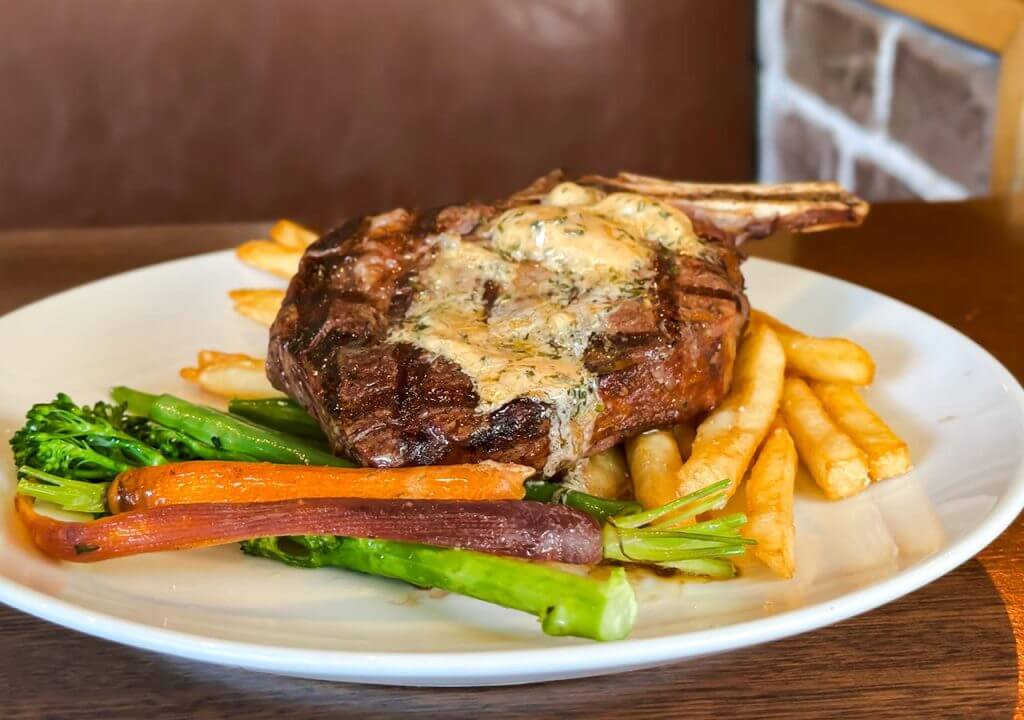 Village Crown Rib Eye Steak Dish containing Cote de Boeuf Steak, Butter, Garlic, Parsley, Thyme, Rosemary, Vegetables, and Fries.